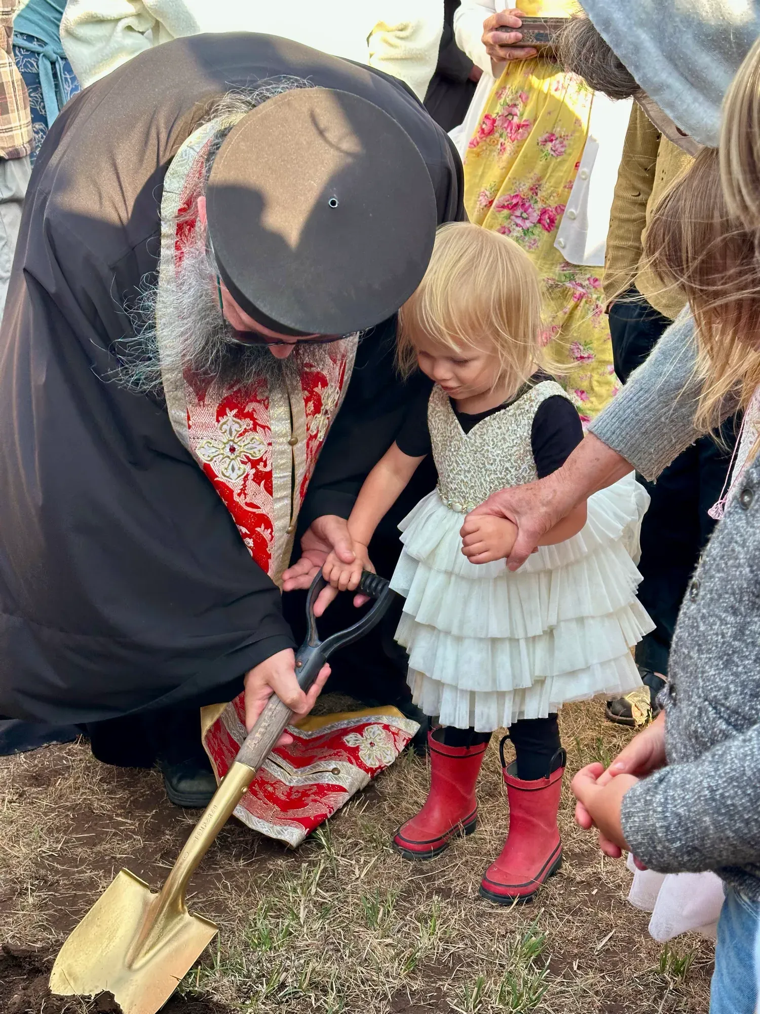 A priest hands a golden shovel to a young child at the groundbreaking