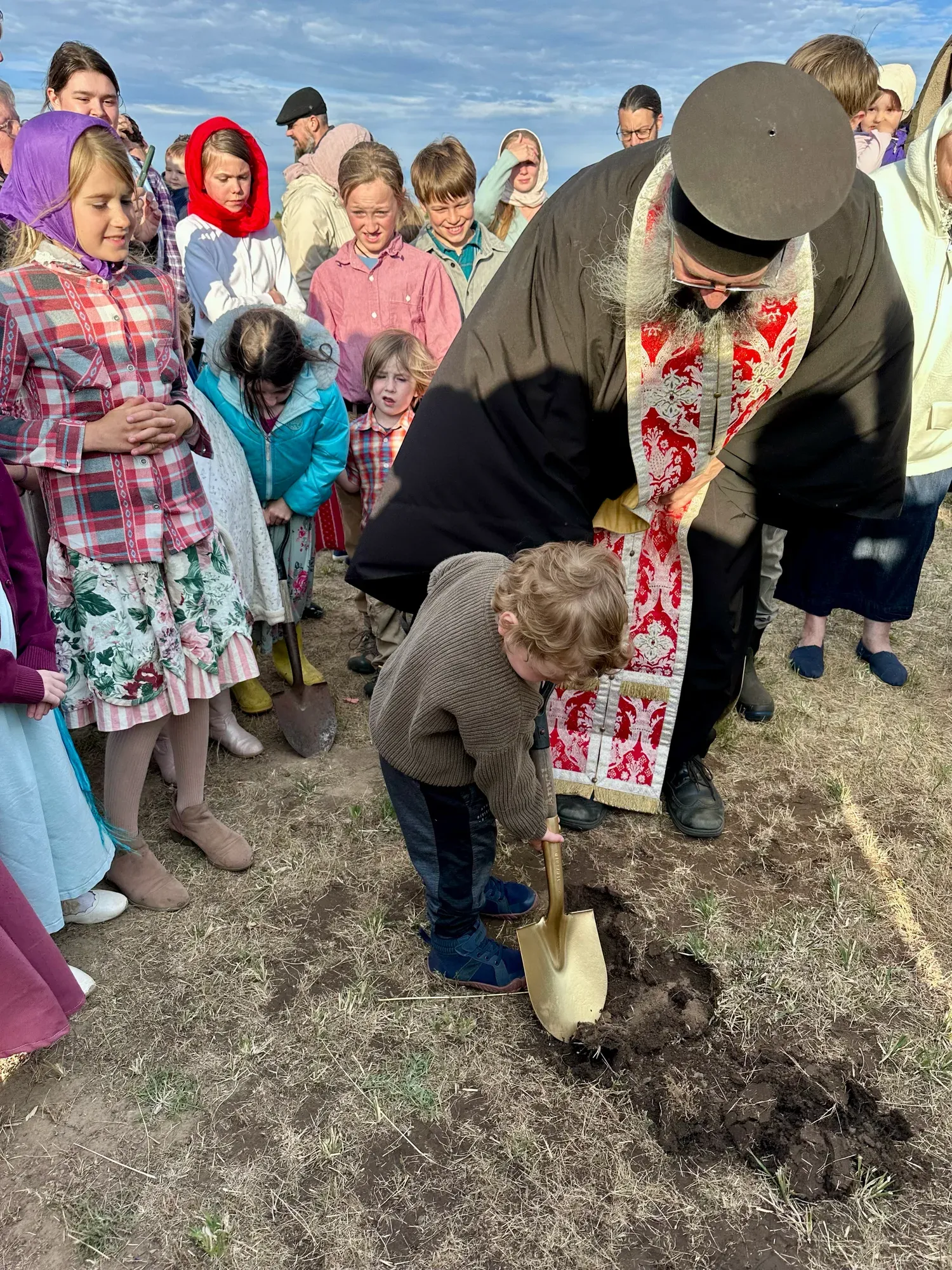 A priest helps a young boy dig with a golden shovel as parishioners look on